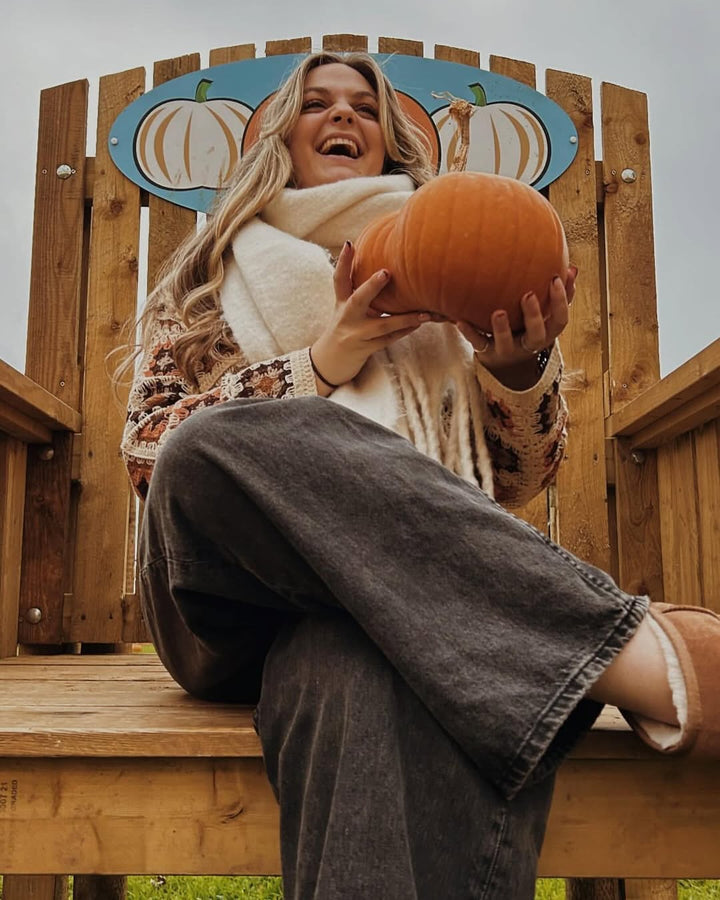 A woman sits on a wooden bench, smiling while holding a pumpkin, with a decorative sign featuring pumpkins behind her.