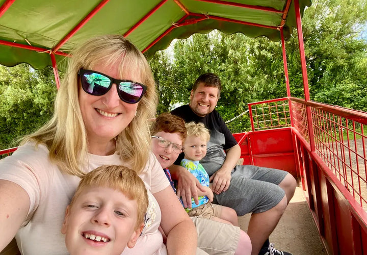 A woman and four children smile together on a red ride, surrounded by greenery on a sunny day.