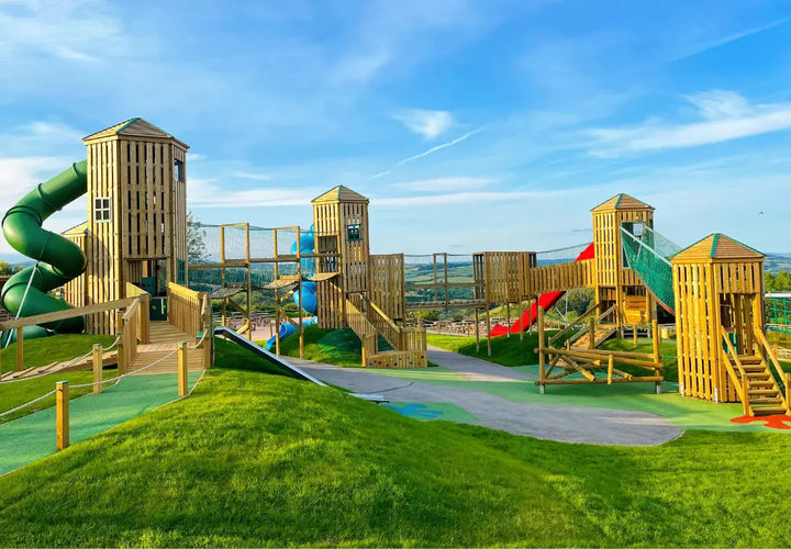 playground with wooden towers, slides, climbing nets, and green grass under a blue sky with scattered clouds