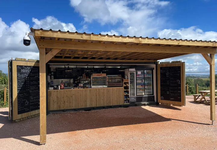 a wooden kiosk with a menu board, serving drinks and snacks, set against a blue sky and green landscape
