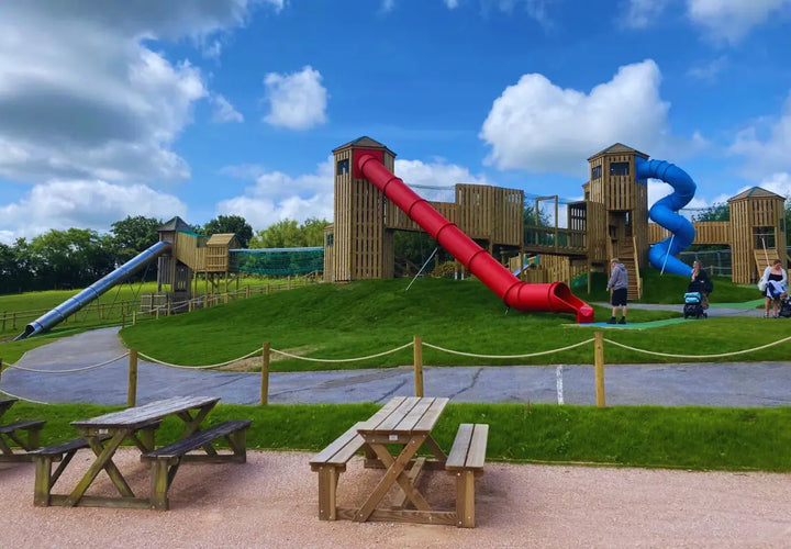 playground with wooden structures, red and blue slides, grassy area, and picnic tables under a blue sky with clouds