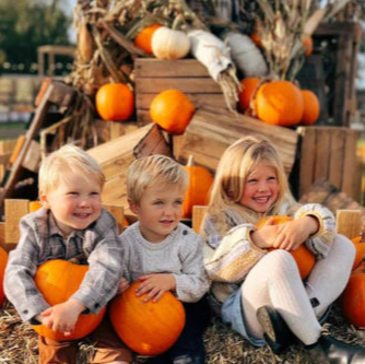 three children sitting on hay bales, each holding a pumpkin, with a backdrop of pumpkins and wooden crates