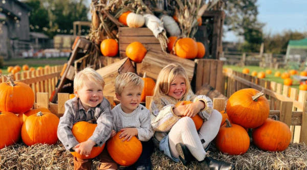 three children sitting on hay bales, holding pumpkins, with a backdrop of pumpkins and a rustic wooden structure